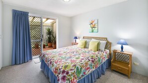 A bedroom with a bed, two bedside tables, a ceiling light, a window with curtains, and a door leading to a balcony with potted plants.