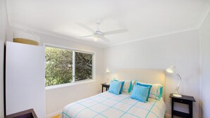 Une chambre à coucher comprenant un lit, deux tables de chevet, un ventilateur de plafond, une fenêtre donnant sur des arbres et un plancher en bois.