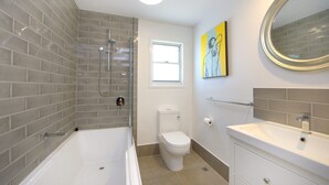 Une salle de bain équipée d’une baignoire, de toilettes et d’un lavabo, avec des murs recouverts de carreaux gris et une fenêtre.