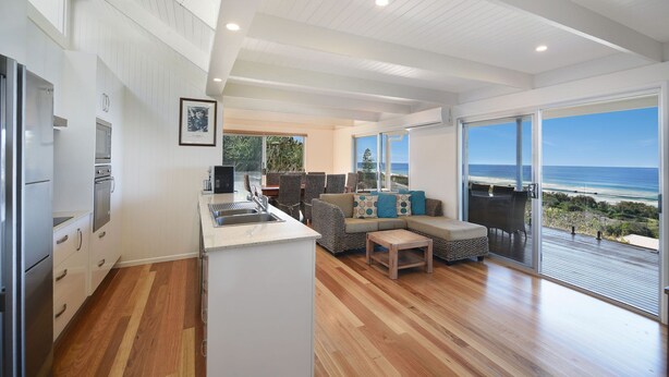 A modern kitchen with white cabinetry and stainless steel appliances, a dining area with a wooden table and chairs, and a large sliding glass door leading to a balcony with a view of the ocean.
