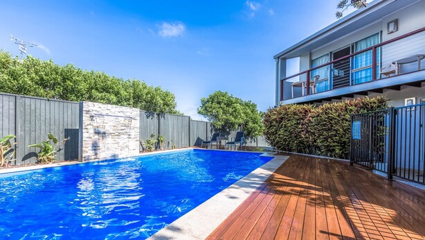A swimming pool with a wooden deck, a stone wall, and a fenced area with greenery. - Lyrona - 3/35 Avocet Parade (Peregian Beach)