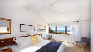 Un dormitorio con una cama grande, un ventilador de techo, una silla y vista al mar a través de las ventanas.