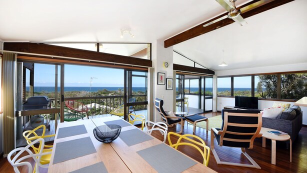 Amplia sala de estar con una gran mesa de madera, sillas coloridas y un sofá. Cuenta con ventanas grandes que ofrecen vistas al océano y un balcón con una parrilla y sillas.