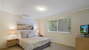 A bedroom with a bed, two bedside tables, a TV, and a window with blinds.