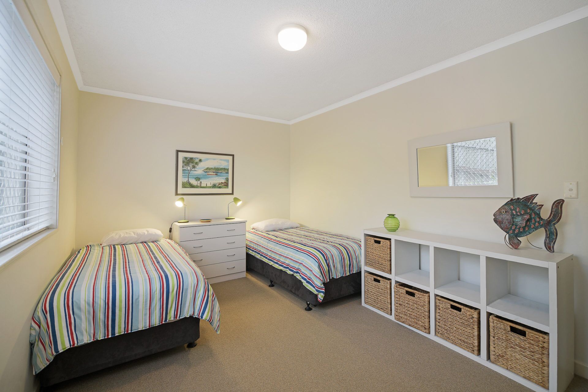 A bedroom with two beds, a dresser, a shelf with baskets, a framed picture, and a ceiling light.
