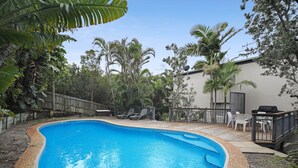 A swimming pool surrounded by a wooden deck, with lounge chairs and a barbecue area.