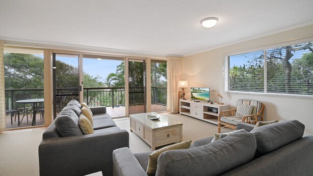A spacious living area with a sofa set, a coffee table, and a TV. There is a balcony with outdoor furniture and a view of greenery.