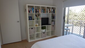 A bedroom with a large white bed, a bookshelf with various items, a television, and a view through a window with a chain-link fence. - Unit 2 at 4 Pelican Street, Peregian Beach, Noosa Area (Peregian Beach)
