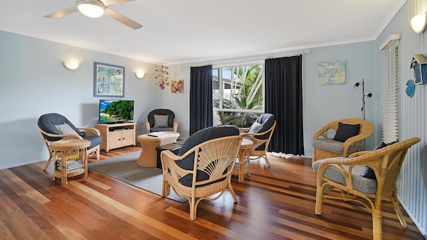 A spacious living room with a ceiling fan, wicker furniture, a television, and a large window with curtains. - Allamanda Sands - 23 Oriole Avenue (Peregian Beach)