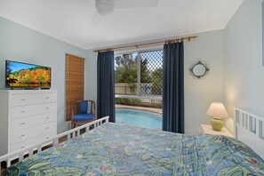 Une chambre avec un lit, une commode, une télévision, une fenêtre avec des rideaux, une lampe et une vue sur une piscine.