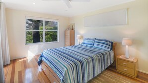 Un dormitorio con cama, mesita de noche, lámpara, ventilador de techo y ventana con vistas a árboles.