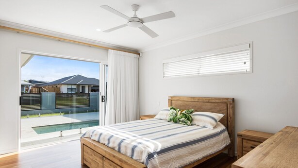 A bedroom with a wooden bed, a ceiling fan, a window with blinds, and a view of a swimming pool and houses outside. - Pet Friendly, new House Peregian Breeze Drive, Peregian Beach (Peregian Beach)