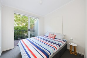 Ein Schlafzimmer mit großem Fenster, einem Bett mit gestreifter Bettwäsche, einem kleinen Nachttisch und Blick ins Grüne.