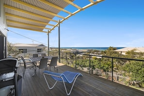 A wooden deck with a table and chairs, overlooking a coastal area with houses and a view of the ocean.