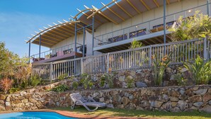 A two-story building with a stone retaining wall, a pool, and a covered outdoor area with a chair. - Peregian Beach House - 2/22 Avocet Parade (Peregian Beach)