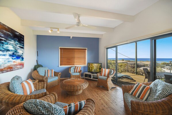 A spacious living room with wicker furniture, a ceiling fan, and a large window offering a view of the ocean.