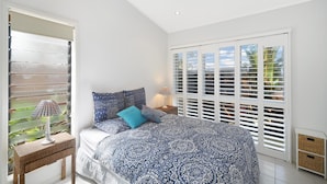 A bedroom with a bed, a bedside table, a lamp, and a window with shutters. - Nobs -1/44 Peregian Esplanade (Peregian Beach)