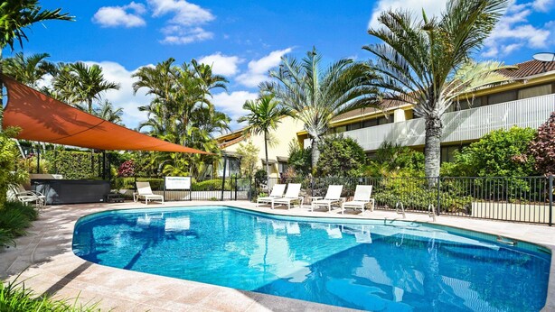 A swimming pool with lounge chairs, surrounded by palm trees and a building with balconies.