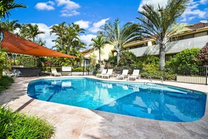 Piscina com espreguiçadeiras, cercada por palmeiras e um prédio com varandas.