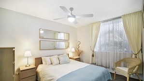 A bedroom with a bed, bedside lamps, a ceiling fan, a mirror, a chair, and a window with curtains.