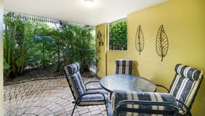 A patio with four striped chairs and a glass table, surrounded by yellow walls and a view of tropical foliage.