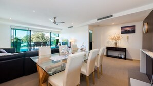 A modern living room with a glass dining table, white chairs, a sofa, and a view of a swimming pool.
