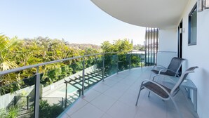 A balcony with a glass railing, two chairs, and a view of a pool and greenery.