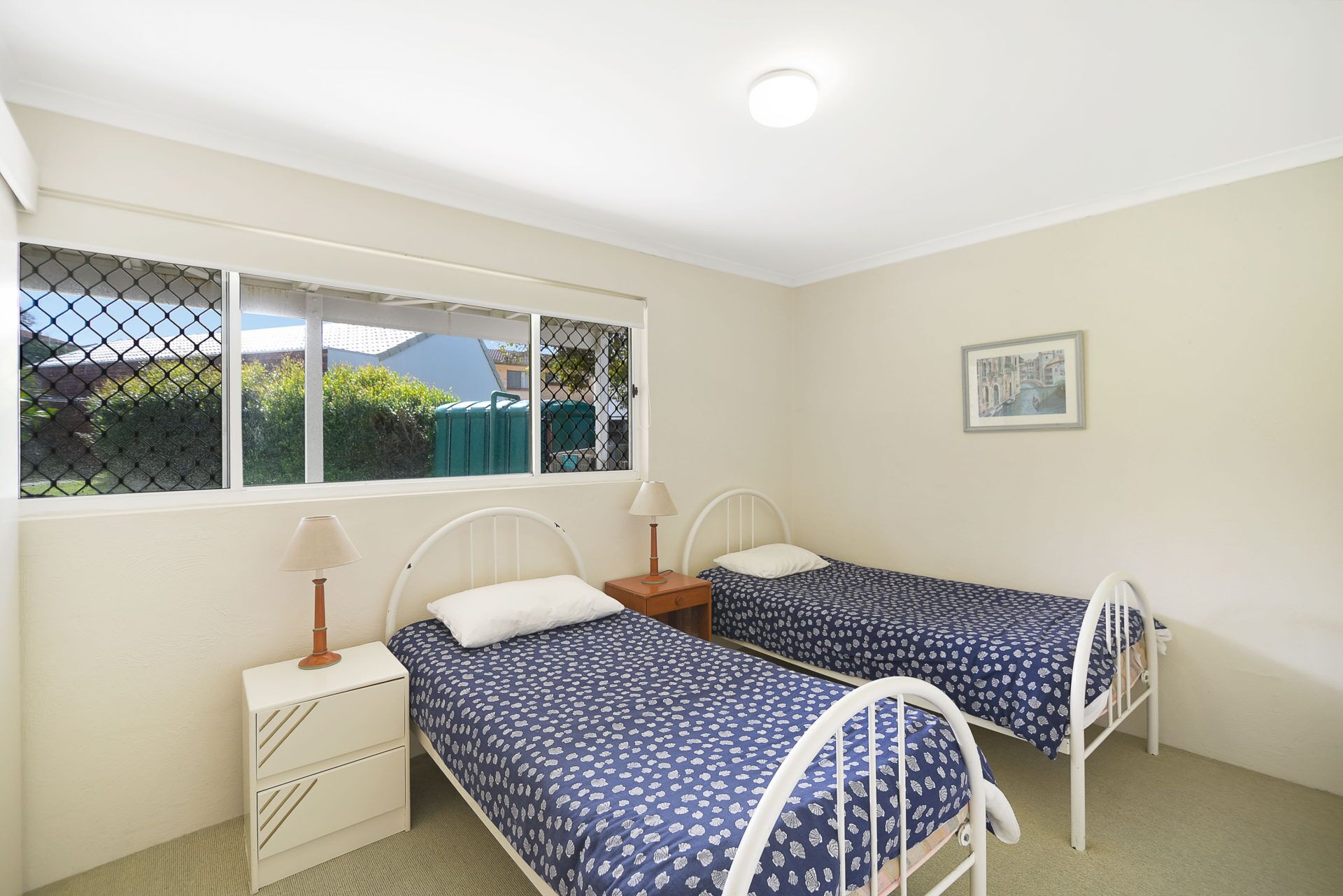 A bedroom with two single beds, a window with a view, a bedside table with a lamp, and a framed picture on the wall.