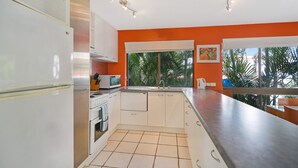 Una cocina con armarios blancos, una estufa y un microondas. Se ve una ventana con vista a palmeras.