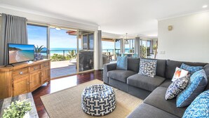 A spacious living room with a large TV, grey sofa, patterned cushions, and a wooden cabinet.