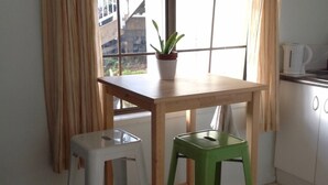 Een kleine keuken met een houten tafel, een groene kruk, een witte kruk, een potplant en een waterkoker op het aanrecht.