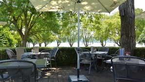 An outdoor patio area with tables and chairs, a large umbrella, and a view of trees and water. - Hotel Iris am See (Radolfzell am Bodensee)