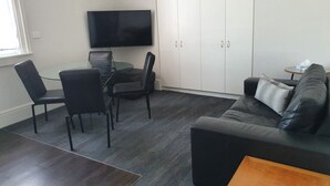 Um espaço compacto com mesa de jantar de vidro, cadeiras pretas, sofá preto, mesa de centro de madeira e televisão fixada na parede.