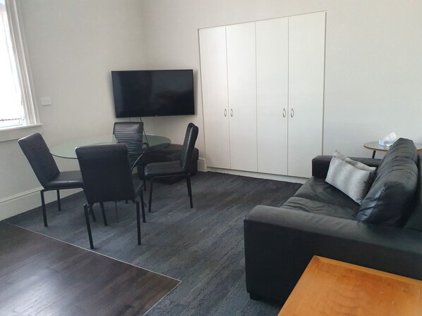 Un espacio habitable compacto con una mesa de comedor de vidrio, sillas negras, un sofá negro, una mesa de centro de madera y un televisor montado en la pared.
