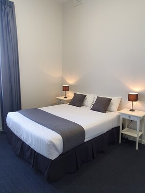 A neatly made bed with a white and gray duvet, two bedside tables with lamps, and a dark blue carpet. - The Parade Hotel (East Bunbury)