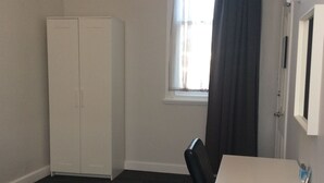 Quarto com guarda-roupa branco, uma secretária pequena com espelho e janela com cortinas.