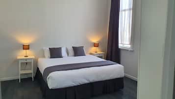 A neatly made bed with a white and dark-colored duvet, two bedside tables with lamps, a window with curtains, and a door.