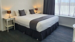 Uma cama bem arrumada com edredom branco e tons escuros, duas luminárias de cabeceira, uma mesinha de cabeceira com gaveta e uma janela com cortina escura.