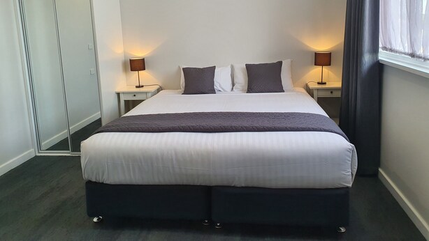 Cama bem arrumada com roupa de cama branca e cinza escura, duas luminárias de cabeceira, um guarda-roupa e uma janela com cortinas.