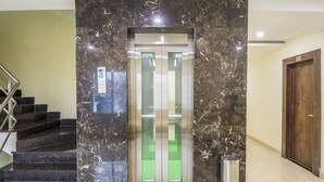 A marble elevator with a glass door, a metal trash can, and a wooden door to the right. - Ostin Viceroy Grand (Guntur)