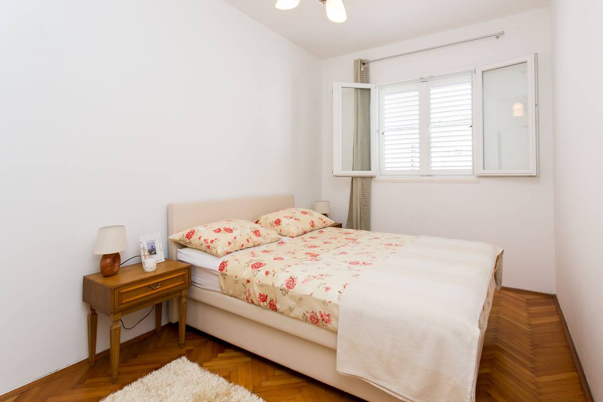 A bedroom with a bed, a nightstand, a lamp, a window with blinds, and a wooden floor.
