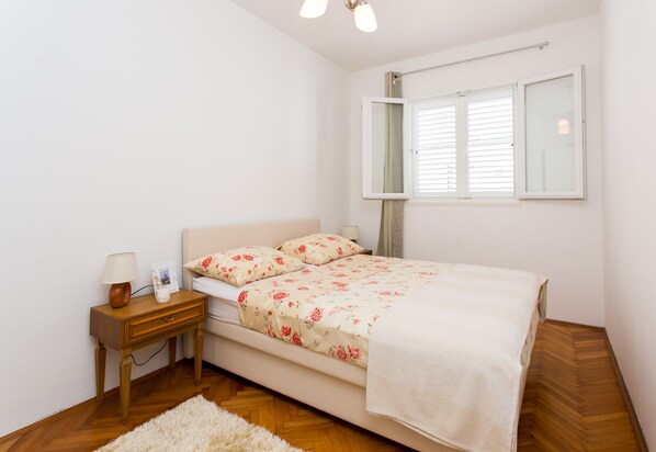 A bedroom with a bed, a nightstand, a lamp, a window with blinds, and a wooden floor. - Studios Heliva (Dubrovnik)