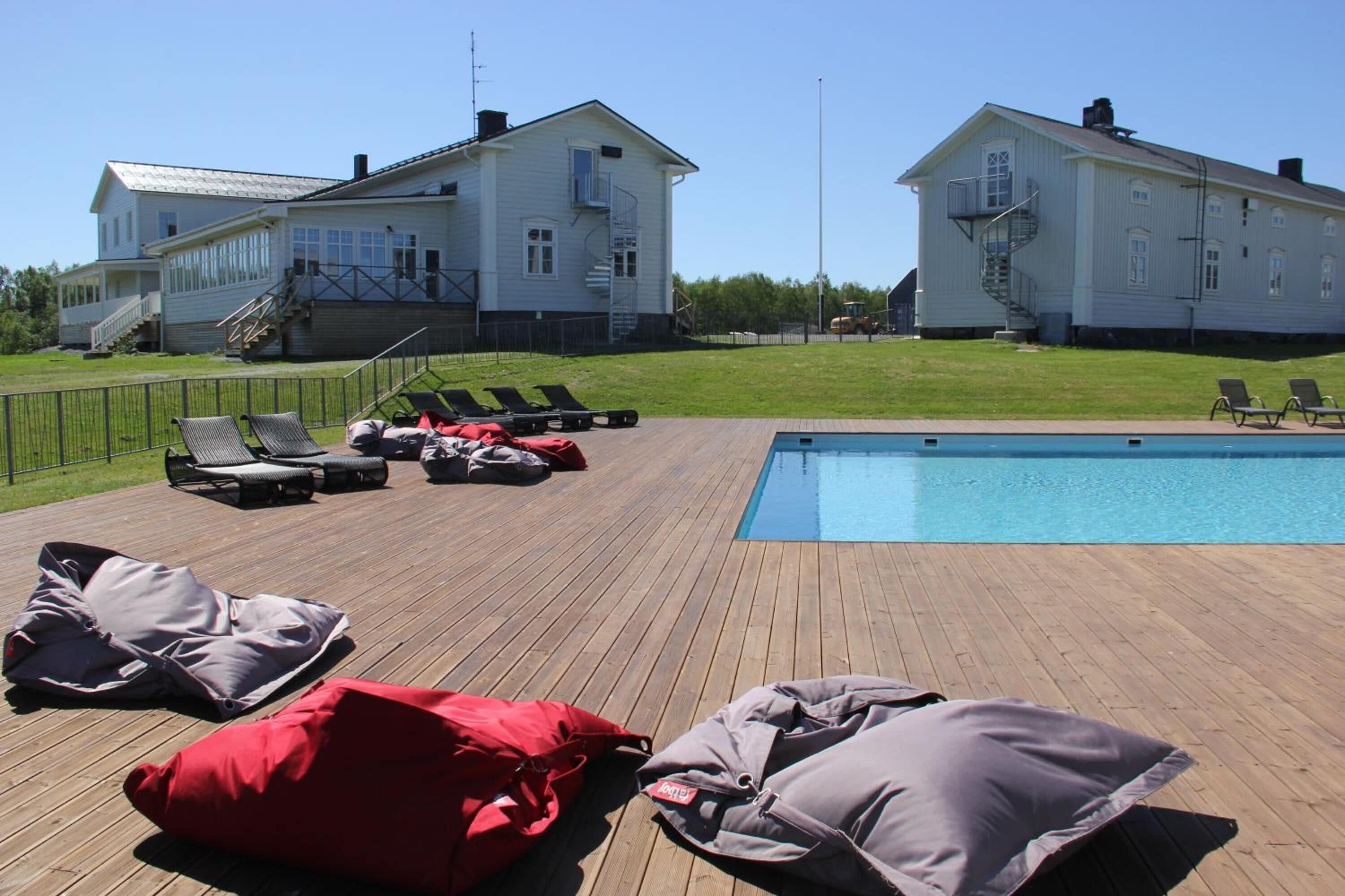 A wooden deck with lounge chairs and a pool, surrounded by two large white houses.