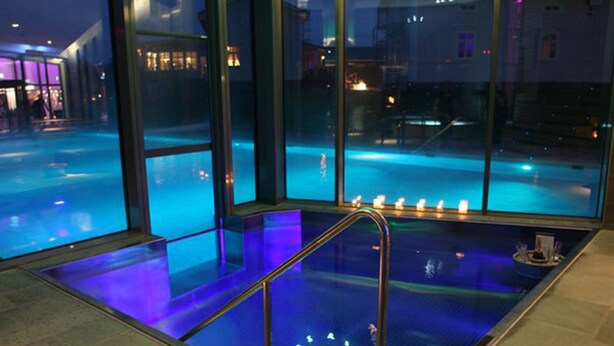 A modern indoor pool with clear glass walls, a blue-lit floor, and a staircase leading down to the pool. - Cape East (Haparanda)