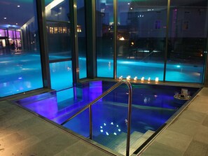 Una piscina interior moderna con paredes de vidrio transparente, un suelo iluminado en azul y una escalera que desciende a la piscina.