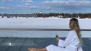 A person sitting on a balcony with a glass table, overlooking a snowy landscape with a bridge and buildings in the distance. - Cape East (Haparanda)