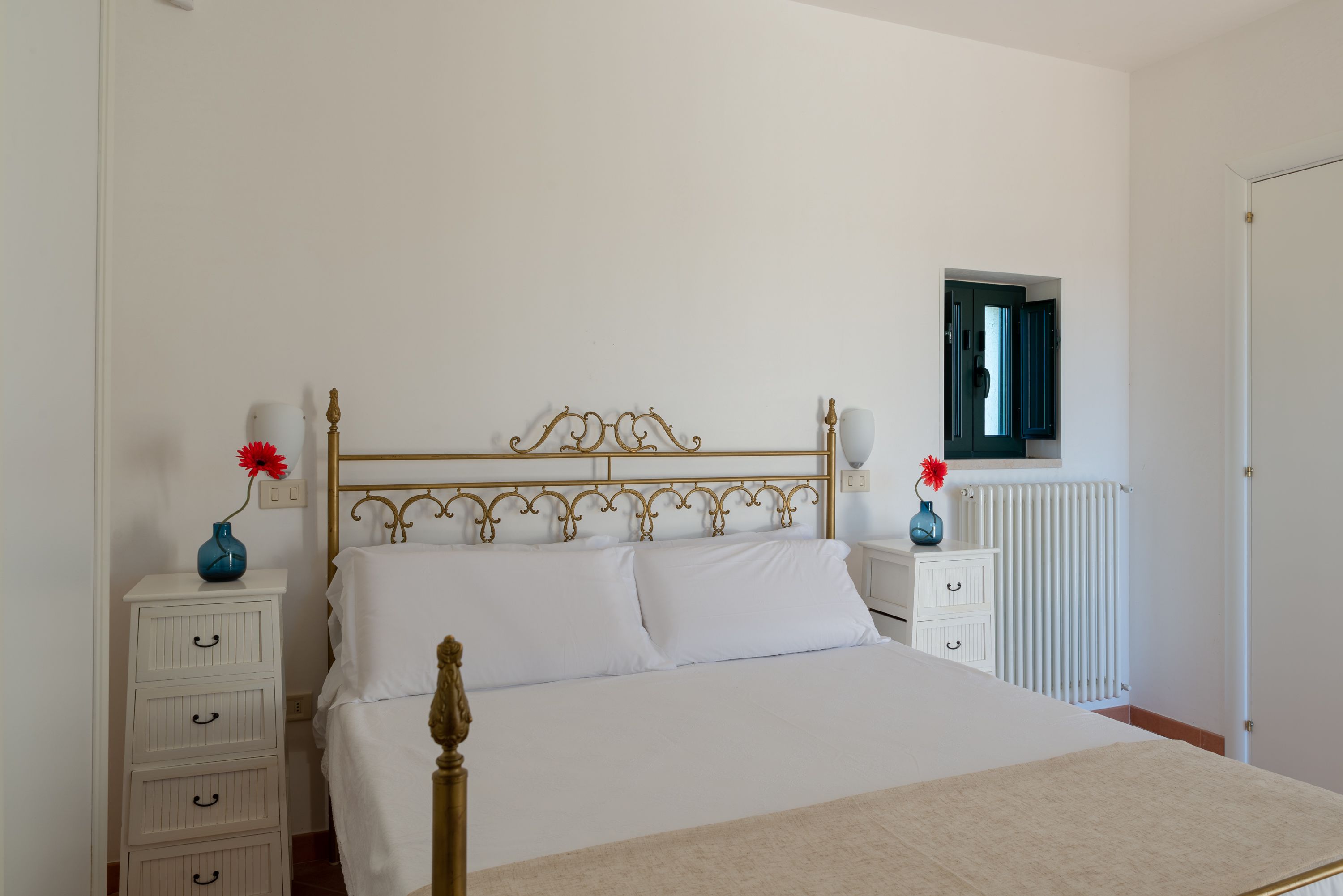A bedroom with a bed, white nightstands, a dresser, a radiator, and a window.