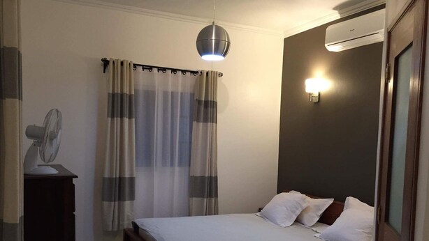 A bedroom with a bed, a bedside table with a fan, a curtain, an air conditioning unit, and a wall-mounted light. - RESIDENCE BAOBAB LOFT (Morondava)