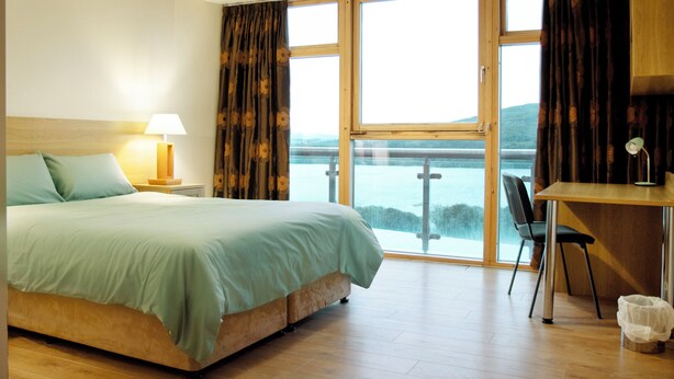A hotel room with a bed, a desk, a chair, and a view of a body of water through large windows.