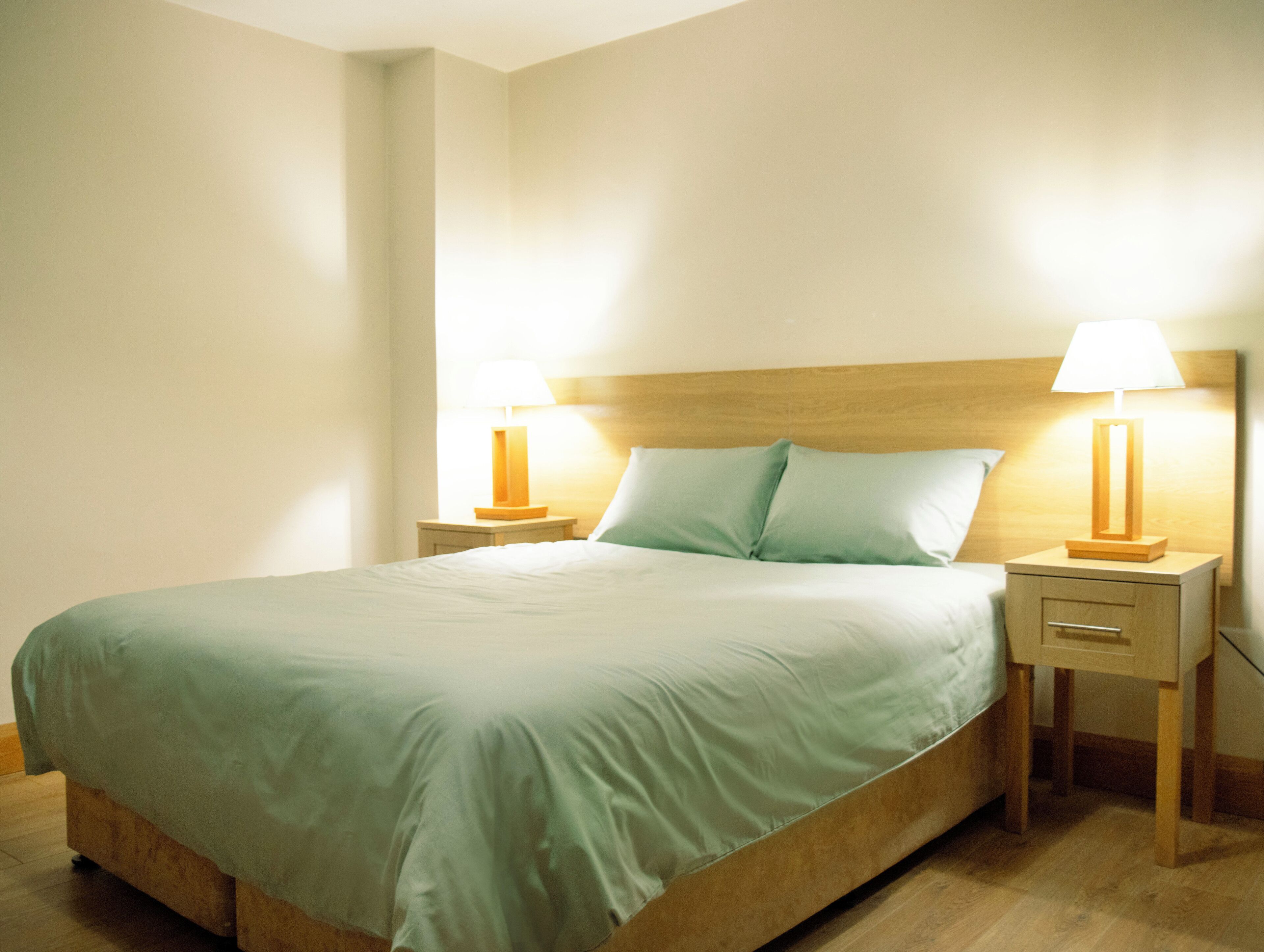 Una cama bien hecha con cabecera de madera, dos mesitas de noche con lámparas y una pared de color claro.